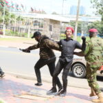 A police officer in camouflage detains a male protester wearing a red beret as a cameraman records the event during a youth-led demonstration against a proposed finance bill.