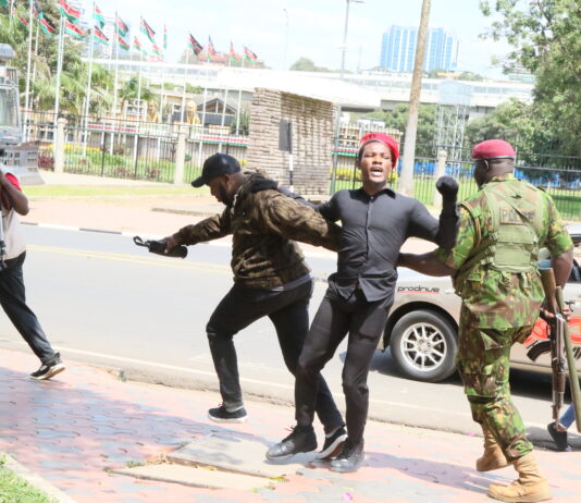 A police officer in camouflage detains a male protester wearing a red beret as a cameraman records the event during a youth-led demonstration against a proposed finance bill.