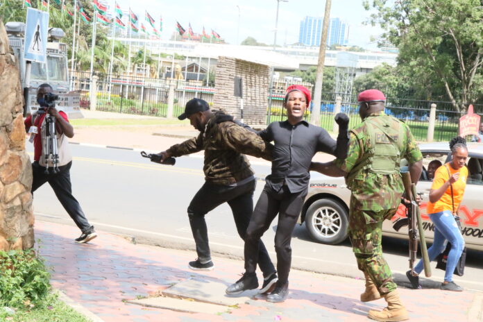 A police officer in camouflage detains a male protester wearing a red beret as a cameraman records the event during a youth-led demonstration against a proposed finance bill.